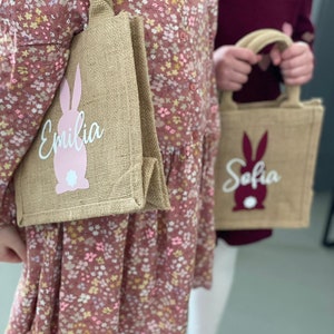 Op de afbeelding: Twee bruine jute tassen met roze konijnenontwerpen en gepersonaliseerde namen. De tas links zegt "Emilia" en de tas rechts zegt "Sofia".