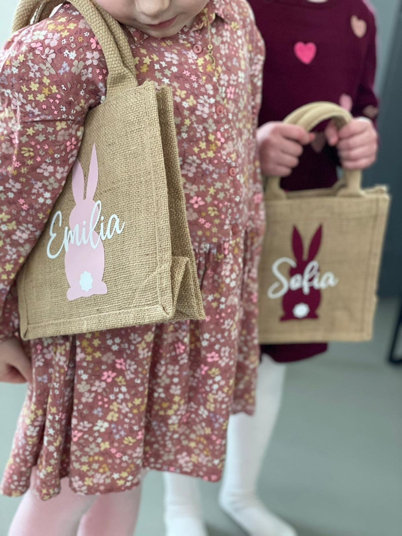 Op de afbeelding: Twee bruine jute tassen met roze konijnenontwerpen en gepersonaliseerde namen. De tas links zegt "Emilia" en de tas rechts zegt "Sofia".