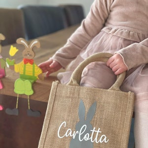 Op de afbeelding: Een bruine jute tas met een grijze konijnen silhouet en de naam "Carlotta" in wit gedrukt. De tas heeft twee bruine handvatten.