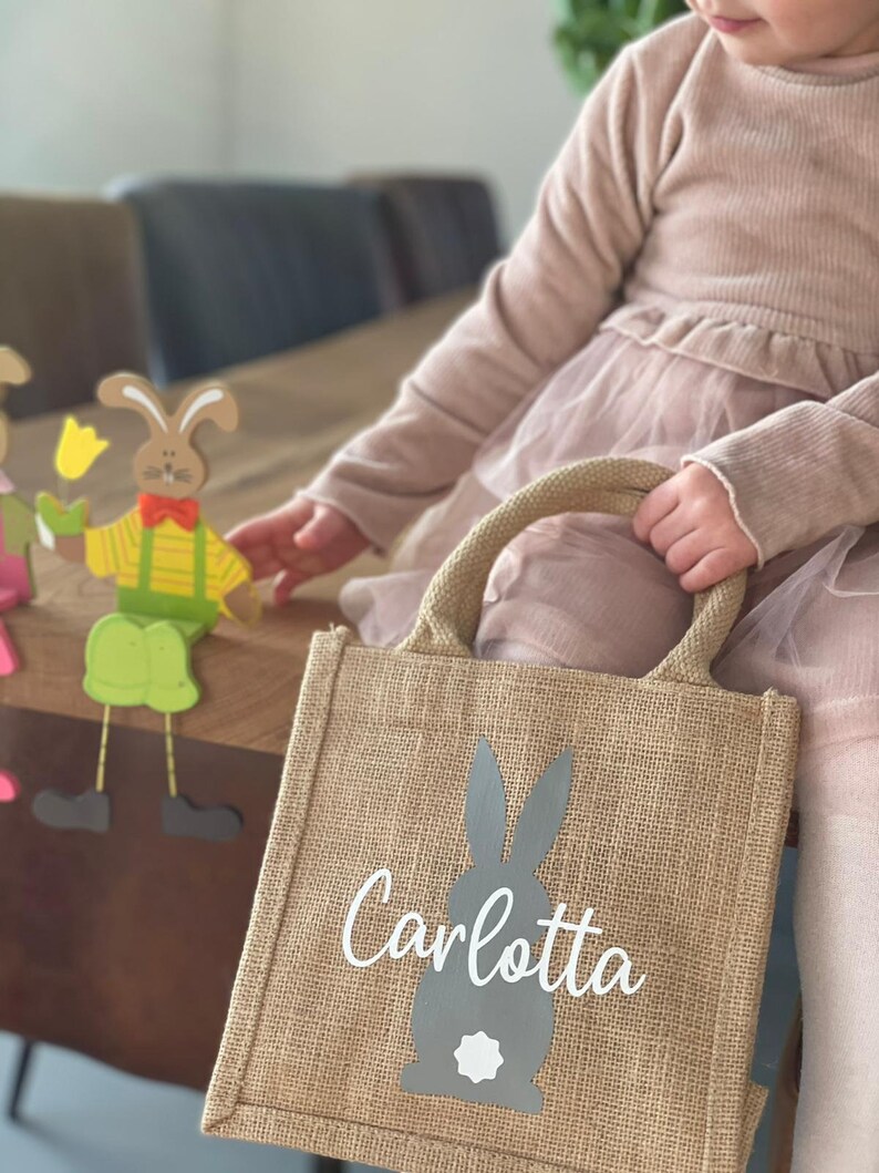 Op de afbeelding: Een bruine jute tas met een grijze konijnen silhouet en de naam "Carlotta" in wit gedrukt. De tas heeft twee bruine handvatten.