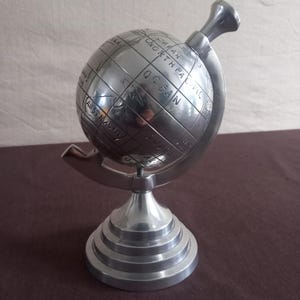 Peut inclure: Une sculpture de globe argenté avec des continents et des océans gravés, montée sur une base à gradins. Le globe est doté d'une poignée sur le dessus et est soutenu par un bras métallique incurvé. Un objet décoratif.