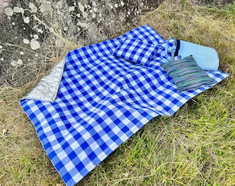 May include: A blue and white checkered picnic blanket with a silver thermal layer on one side. The blanket is folded and laid out on green grass with two pillows on top.