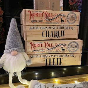 May include: A stack of wooden Christmas Eve boxes, each with the text "North Pole Trading Company" and "Special Christmas Eve Delivery For" followed by names. A plush gnome with a gray hat and white beard is in the foreground. The boxes have red and black text.