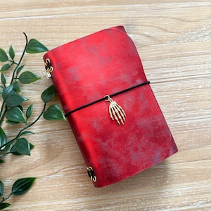 May include: A red leather journal with a black elastic closure and a gold-colored skeleton hand charm. The journal has a distressed finish and gold-colored corner protectors. The background is a light wood surface.