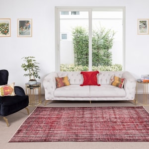 May include: A white sofa with red, yellow, and orange throw pillows sits in a living room with a black armchair and a red rug. The room has a large window with a view of green trees and a white wall with framed artwork.