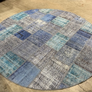 Może przedstawiać: Okrągły dywan typu patchwork składający się z różnych kwadratów tkaniny w odcieniach niebieskiego, turkusowego i szarego. Każdy kwadrat posiada wyraźne, wyblakłe wzory i tekstury, zszyte razem w okrągłą wykładzinę z widocznym szwem na krawędzi.