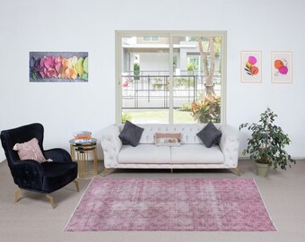 May include: A white sofa with two grey throw pillows and a patterned throw pillow sits in a living room with a black armchair, a side table with books, and a pink rug. The room has a large window with a view of a garden and two framed prints on the wall.