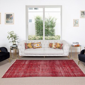 Könnte beinhalten: Ein Wohnzimmer mit einem hellgrauen, getufteten Sofa, zwei schwarzen Samtsesseln und einem roten, abgenutzten Teppich. Dekorative Kissen mit geometrischen Mustern und gerahmte Kunstwerke schmücken die Wände. Kleine goldene Beistelltische und eine Topfpflanze vervollständigen die Einrichtung.