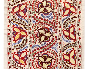 May include: A white cotton fabric with a repeating pattern of red, yellow, blue, and green embroidered flowers. The flowers are stylized and have a central point with four petals. The background is a cream color.