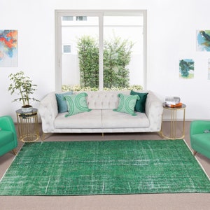 May include: A living room with a white sofa, two green armchairs, and a green rug. The sofa has two green pillows and a white throw pillow. The armchairs have a pink throw pillow. There are two gold side tables with books and a plant on one of them. There is a window with a view of trees and a white wall with three abstract paintings.