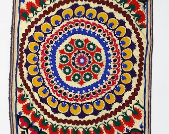 May include: A colorful embroidered textile with a central circular design featuring concentric circles of yellow, blue, red, and green. The textile is surrounded by a border of floral motifs in shades of pink, red, green, and blue.