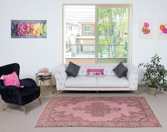 May include: A living room with a light pink rug featuring a central medallion design. A white sofa with grey cushions and a black armchair with a pink cushion are visible. Artwork and a potted plant add decorative elements.