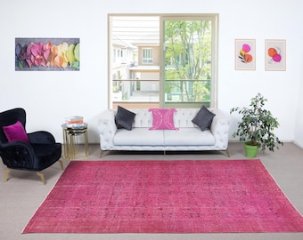May include: A living room scene featuring a large, rectangular, distressed fuchsia rug. A white tufted sofa with gray and pink pillows sits in front of a window. A black armchair with a purple pillow and a gold side table complete the decor.