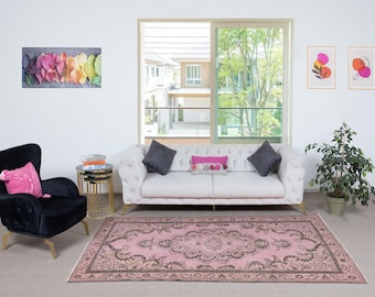 May include: A living room with a pink and brown patterned rug, a white tufted sofa with grey cushions, and a black armchair with a pink cushion. Artwork and a potted plant complete the scene.