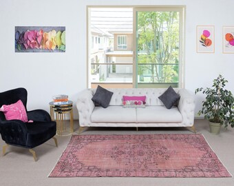 May include: A living room with a light pink vintage rug with a dark border. A white tufted sofa with grey cushions is against a window. A black armchair with a pink cushion is on the left. Artwork and a small plant complete the decor.