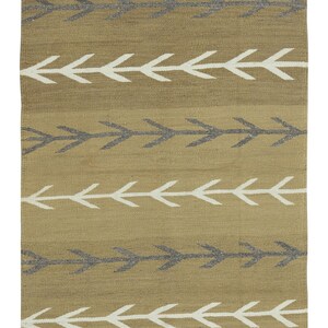 Puede incluir: Una alfombra rectangular con un patrón de flechas repetidas. La alfombra presenta filas de flechas blancas y grises sobre un fondo beige. El borde tiene rayas negras, blancas y grises.