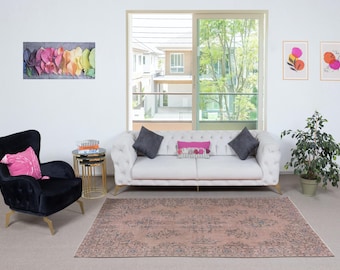 May include: A living room with a white sofa, grey and pink cushions, a black armchair, and a patterned rug. Artwork and a potted plant are also visible. The room is bright and airy, with a view through a window.
