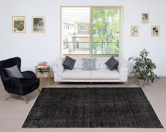 May include: A living room with a dark, patterned rug with a white edge. A white, button-backed sofa with grey cushions is in front of a window. A black armchair and a small side table are also visible, along with framed pictures.