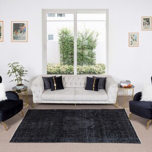 May include: A living room interior design with a white sofa, two black velvet armchairs with gold legs, and a black and white patterned rug. The sofa has two black and gold pillows. There are two gold side tables with glass tops and a small potted plant.