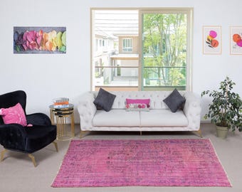 Könnte beinhalten: Ein Wohnzimmer mit einem weißen, getufteten Sofa, grauen Kissen und einem rosa gemusterten Kissen. Links steht ein schwarzer Sessel mit einem rosa Kissen. Ein großer rosa Teppich bedeckt den Boden. Kunstwerke und Pflanzen ergänzen die Einrichtung.