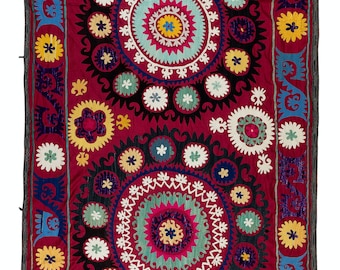 May include: A vibrant red textile with intricate embroidery featuring colorful floral motifs and geometric patterns. The design includes multiple concentric circles with a central starburst and a border of stylized leaves.