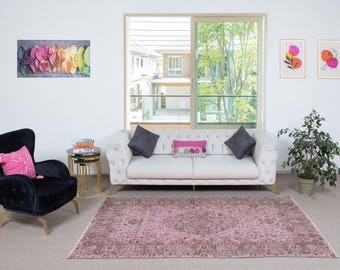 May include: A living room with a white sofa, grey and pink cushions, and a black armchair. A pink patterned rug covers the floor. Artwork and a plant are present. A window provides a view of the exterior.