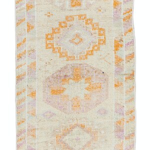 Könnte beinhalten: Ein Vintage-Läuferteppich mit cremefarbenem Hintergrund und einem geometrischen Muster in Orange- und Rosatönen. Der Teppich hat einen Fransenrand.