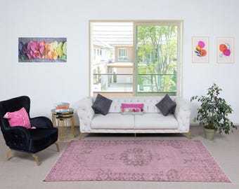 May include: A living room with a light pink rug featuring a central medallion design. A white sofa with grey cushions is near a window. A black armchair with a pink cushion is on the left. Artwork and a potted plant complete the decor.