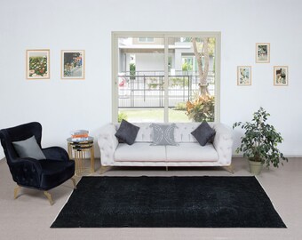 Pode incluir: Um sofá branco com almofadas cinzas fica em uma sala de estar com um tapete preto, uma poltrona preta e uma mesa lateral dourada. Há fotos emolduradas na parede e uma planta em vaso no canto.