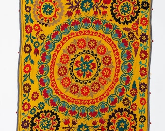 May include: A yellow fabric square with intricate embroidery in red, blue, green, and black. The design features a central circle surrounded by smaller circles and floral motifs.