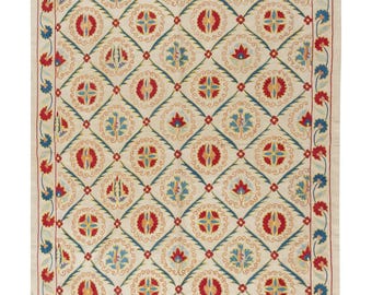 May include: Cream-colored textile with a repeating geometric pattern of red, blue, and yellow floral motifs. The design is framed by a border of red and blue flowers. The textile is rectangular and appears to be a rug or wall hanging.