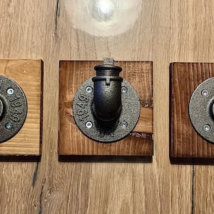 Könnte beinhalten: Drei schwarze Metall-Rohrhaken, die auf quadratischen Holzblöcken montiert sind. Jeder Haken hat eine runde Basis mit der Nummer "1949" darauf.