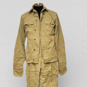 May include: A tan military uniform comprising a button-down jacket with two chest pockets and trousers with stirrups. The uniform is displayed on a mannequin against a plain white backdrop.
