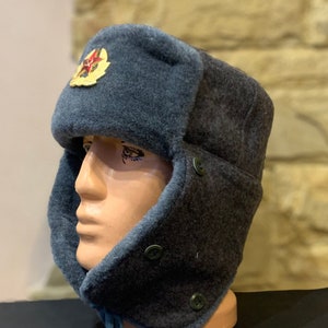 May include: A dark blue and grey winter hat with ear flaps and a red star emblem. The hat has a button fastening on the side.