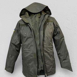 Może przedstawiać: Zielona kurtka w stylu wojskowym z kapturem. Kurtka posiada liczne kieszenie, zapięcie na zamek błyskawiczny oraz naszywkę z niebiesko-żółtą flagą. Zaprojektowana do użytku na zewnątrz i ochrony przed warunkami atmosferycznymi.