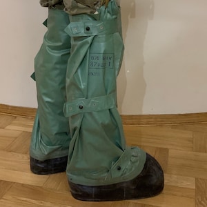 Puede incluir: Un par de polainas impermeables de estilo militar verde con correas y hebillas negras. Las polainas se llevan sobre las botas y tienen una suela de goma negra. El texto "076 BLK 37 P001 NTK 20E" está impreso en el lateral de las polainas.