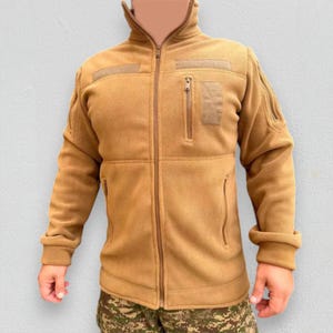 May include: A tan fleece jacket with a full-length zipper, two front pockets, and a high collar. The jacket has two square patches on the chest and a small zippered pocket. The sleeves have additional zippered pockets. The jacket is shown with camouflage pants.