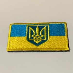 May include: A yellow and blue embroidered patch featuring the Ukrainian coat of arms, a trident, on a yellow background with a blue stripe above it.
