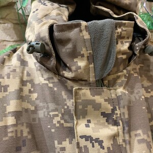 May include: Close-up of a camouflage jacket with a zipper and a hood. The jacket has a drawstring at the hood and a small black toggle on each side of the hood.