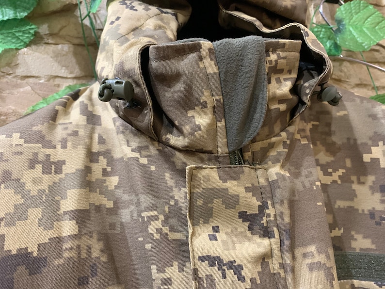 May include: Close-up of a camouflage jacket with a zipper and a hood. The jacket has a drawstring at the hood and a small black toggle on each side of the hood.