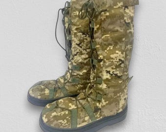 May include: A pair of tall, calf-length boots featuring a digital camouflage pattern in shades of olive green, tan, and brown. The boots have a lace-up front closure with matching fabric loops and a solid, dark gray rubber sole and toe cap.