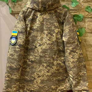 May include: A camouflage jacket with a hood, featuring a Ukrainian flag patch on the right sleeve and a military patch on the left sleeve. The jacket has a zipper closure and multiple pockets.