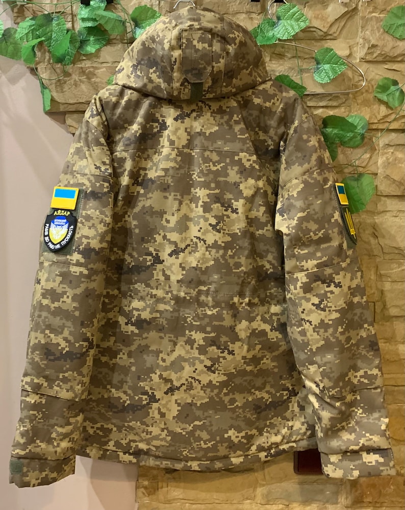 May include: A camouflage jacket with a hood, featuring a Ukrainian flag patch on the right sleeve and a military patch on the left sleeve. The jacket has a zipper closure and multiple pockets.