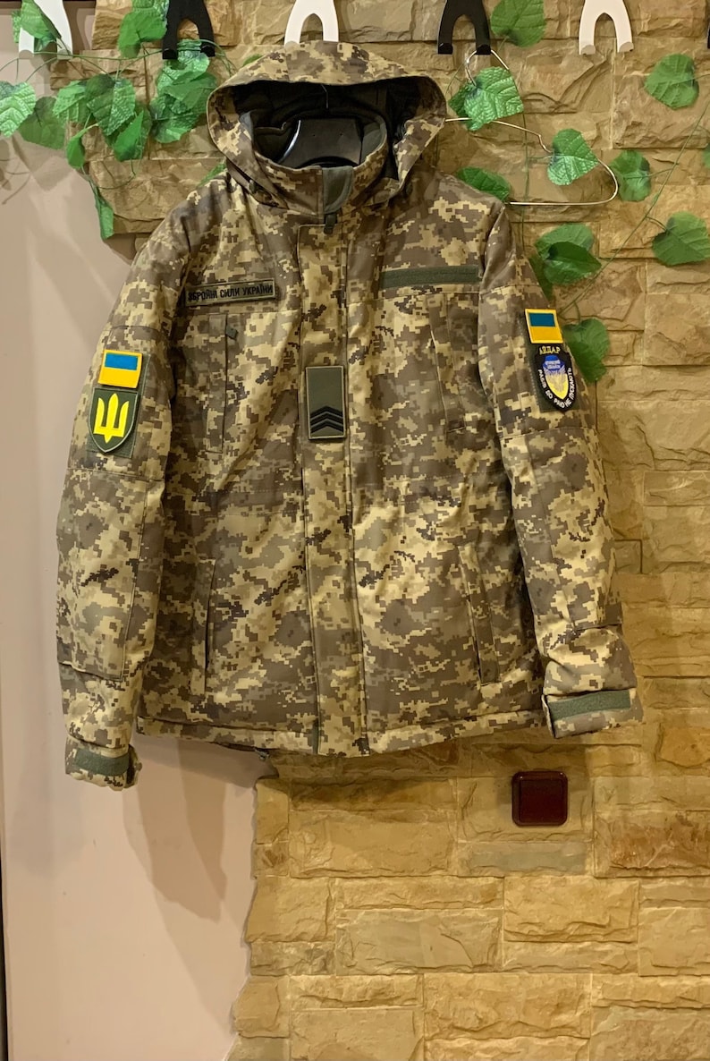 Ukrainian winter field jacket in Pixel MM-14 camouflage hanging on hanger, front view, hood visible, natural indoor lighting.