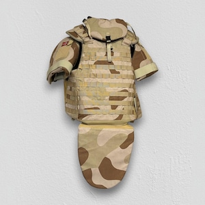 May include: A desert camouflage tactical vest featuring a modular webbing system, shoulder guards, and a detachable groin protector. The vest is constructed with tan, beige, and brown patterns, including a small red patch on the right shoulder.