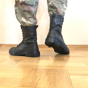 May include: A pair of black leather combat boots with laces. The boots have a thick sole and are worn by a person wearing camouflage pants.