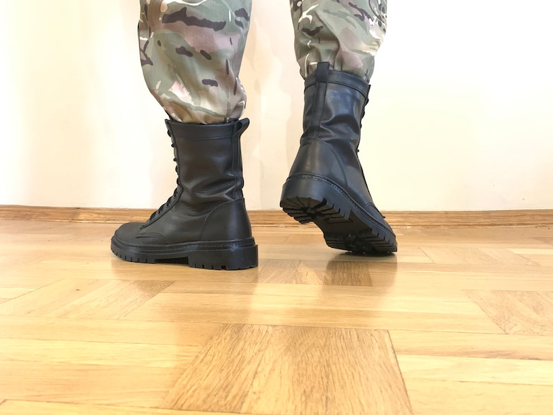 May include: A pair of black leather combat boots with laces. The boots have a thick sole and are worn by a person wearing camouflage pants.