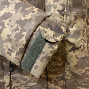 May include: Close-up of a camouflage jacket sleeve with a green velcro strap. The fabric is a pixelated pattern of brown, tan, and green.