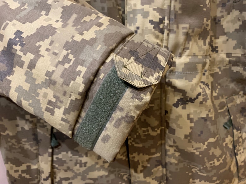 May include: Close-up of a camouflage jacket sleeve with a green velcro strap. The fabric is a pixelated pattern of brown, tan, and green.
