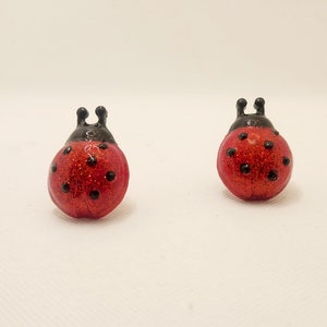 May include: Two ladybug figurines. Each ladybug has a red, glittery body with black spots and a black head with antennae. The figurines are set against a white background.